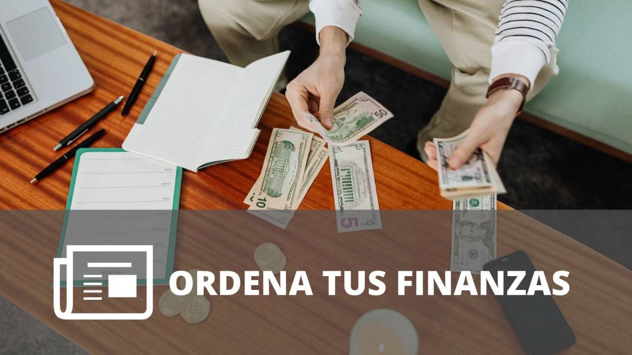 Persona organizando dinero en efectivo y revisando sus finanzas sobre una mesa con libreta, computadora y teléfono. 