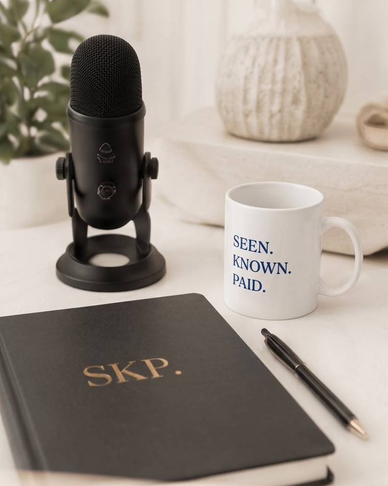 Podcast microphone beside an SKP notebook and Seen Known Paid mug