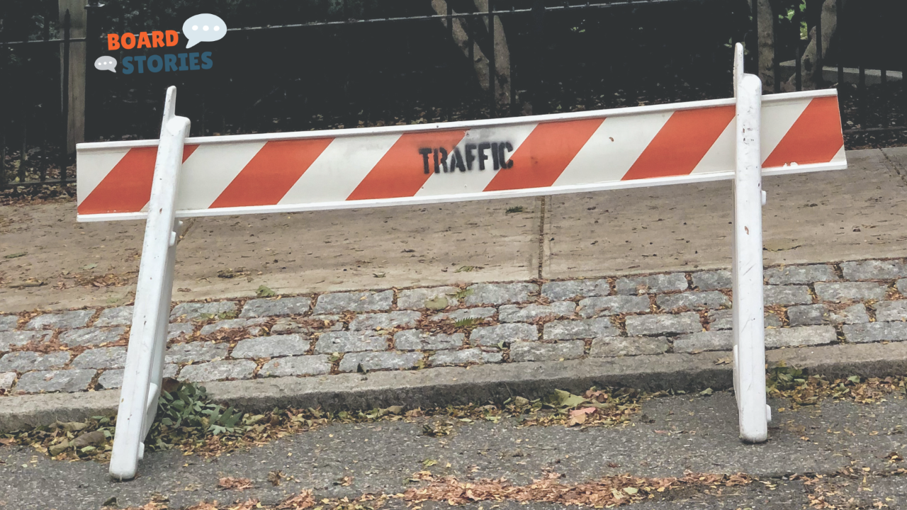 A white and orange striped traffic barricade stamped with the word "TRAFFIC" stands on a cobblestone sidewalk beside a wrought iron fence, with fallen autumn leaves scattered on the ground. The Board Stories logo appears in the upper left corner.