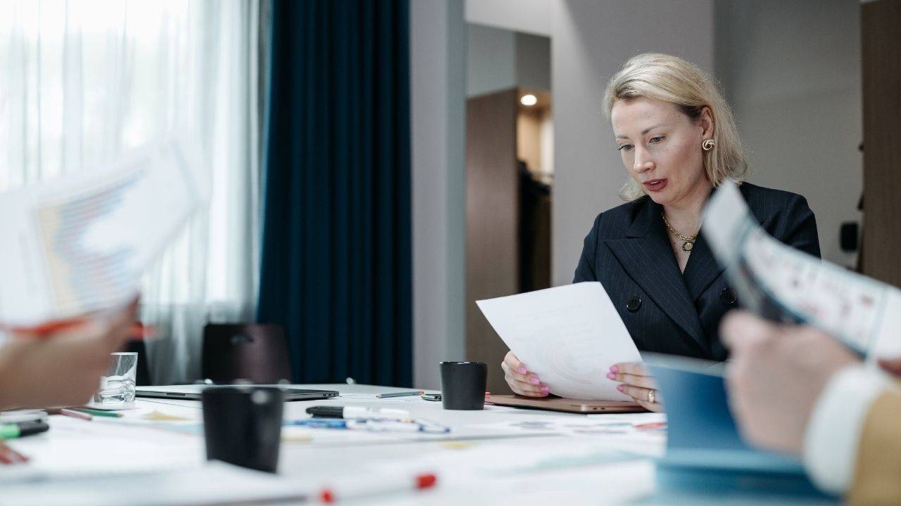 Senior woman leader reviews reports at a conference table while colleagues present documents, illustrating team decision-making concentrated with one leader.
