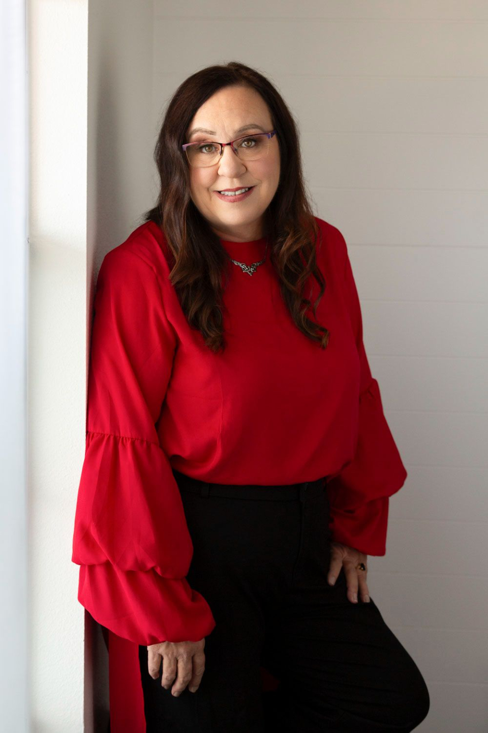 Amanda L. Christian, author of SoulFIRE Leadership, long dark hair, glasses, wearing a red ruffled sleeve blouse, leaning casually against a light gray wall, executive coach and leadership strategist for high achieving women in male dominated workplaces.