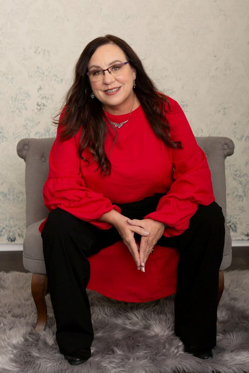 Amanda L Christian, a Master Life Coach and thought leader in burnout recovery and leadership development, sits confidently on a stylish gray chair. Dressed in a bold red blouse with statement sleeves and black trousers, she exudes warmth, wisdom, and approachability. Her long brunette hair cascades over her shoulders, and she wears glasses, a silver necklace, and a genuine smile, creating a professional yet inviting presence against a soft, elegant backdrop.