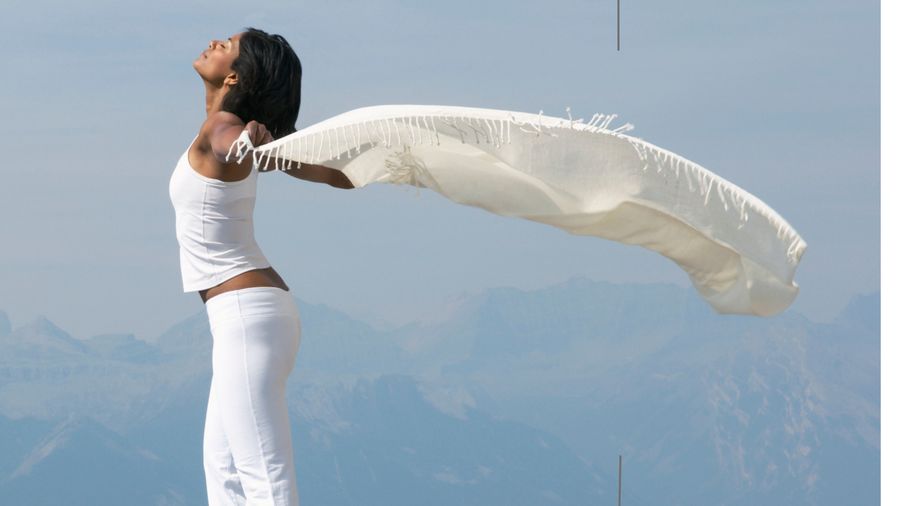 Woman in white standing against a mountain sky, a long scarf blowing in the wind
