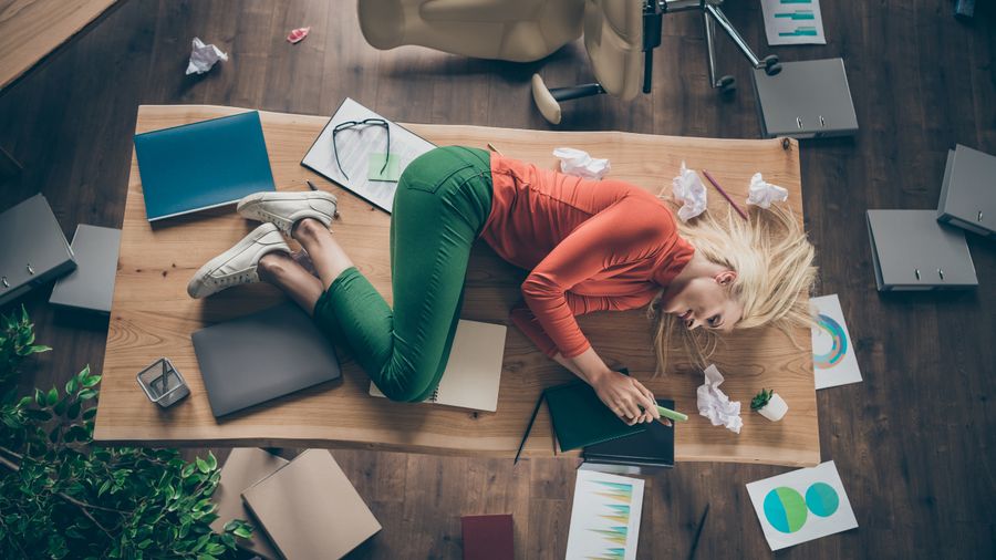 Woman lying across a cluttered desk surrounded by papers and folders, seen from above