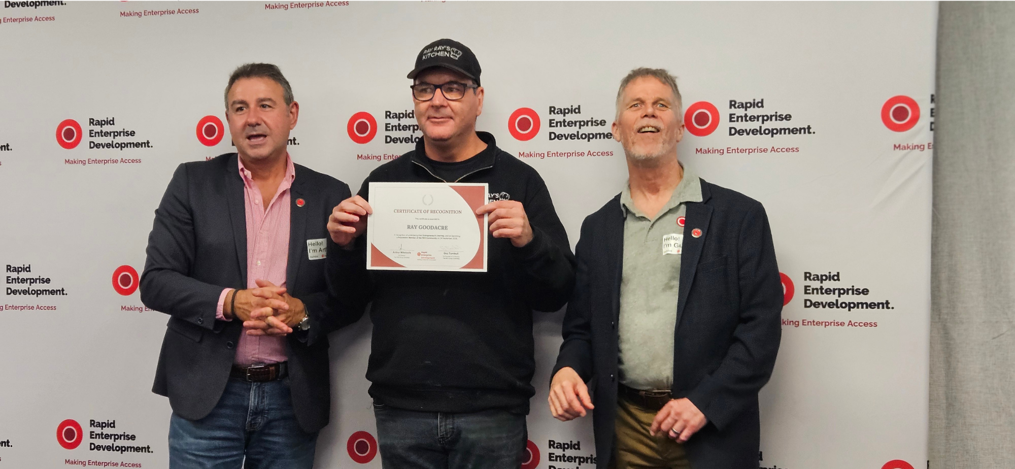 Three people stand before a Rapid Enterprise Development backdrop; the center person holds a framed certificate awarded to Ray Goodacre.