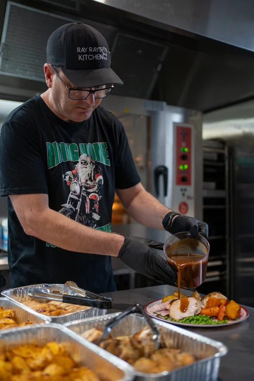 Man in a commercial kitchen wearing a 'Ray Ray’s Kitchen' cap and King Kong T-shirt, plating food with gravy over meat, vegetables, and roasted potatoes beside trays of cooked dishes.