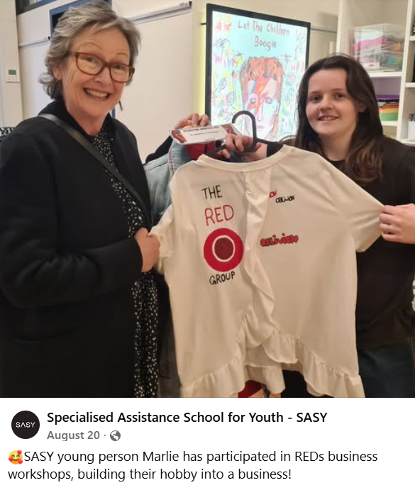 Two people hold a RED Group ambassador shirt in a classroom with 'Let The Children Boogie' artwork