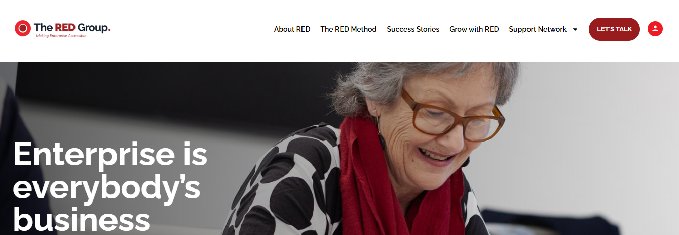 image Webpage header for The RED Group featuring the logo with a red circle and text ‘The RED Group. Making Enterprise Accessible.’ The navigation bar includes links for About RED, The RED Method, Success Stories, Grow with RED, Support Network, and a red ‘LET’S TALK’ button. Below, a smiling older woman with gray hair, glasses, and a red scarf is reading or writing at a desk beside large white text that says ‘Enterprise is everybody’s business.