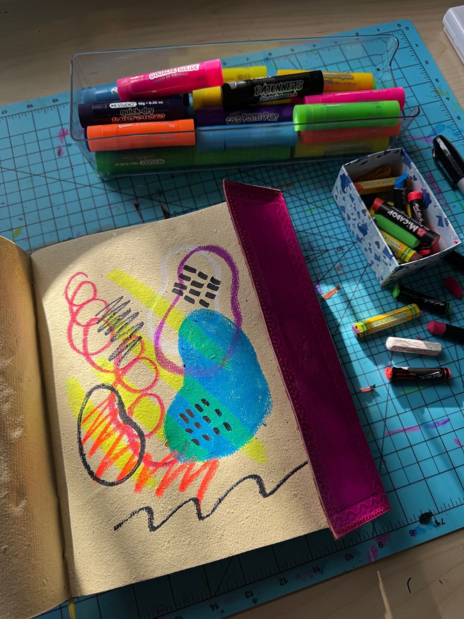 A sketchbook page of bright oil pastel shapes next to a tray of paint sticks and oil pastels on a cutting mat