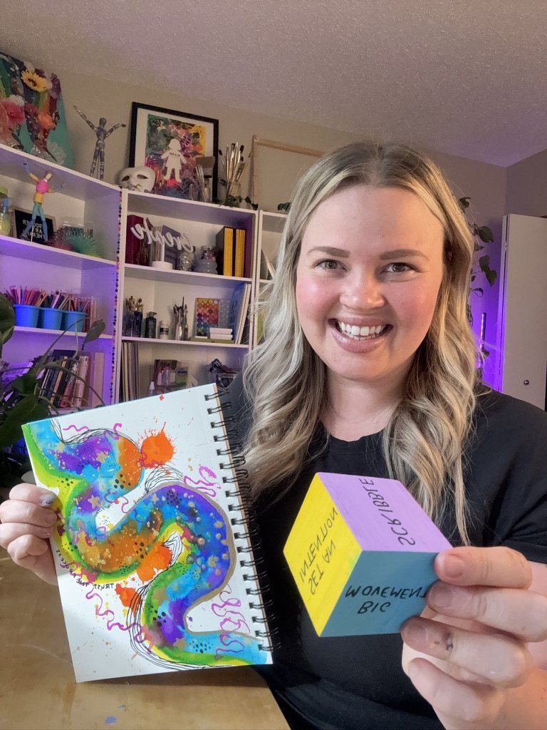 Ashleigh smiling at her art shelves, holding her sketchbook and a painted cube