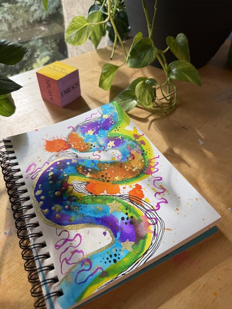 A watercolour page of rainbow swirls with gold stars in a spiral sketchbook, with a painted cube and a plant beside it