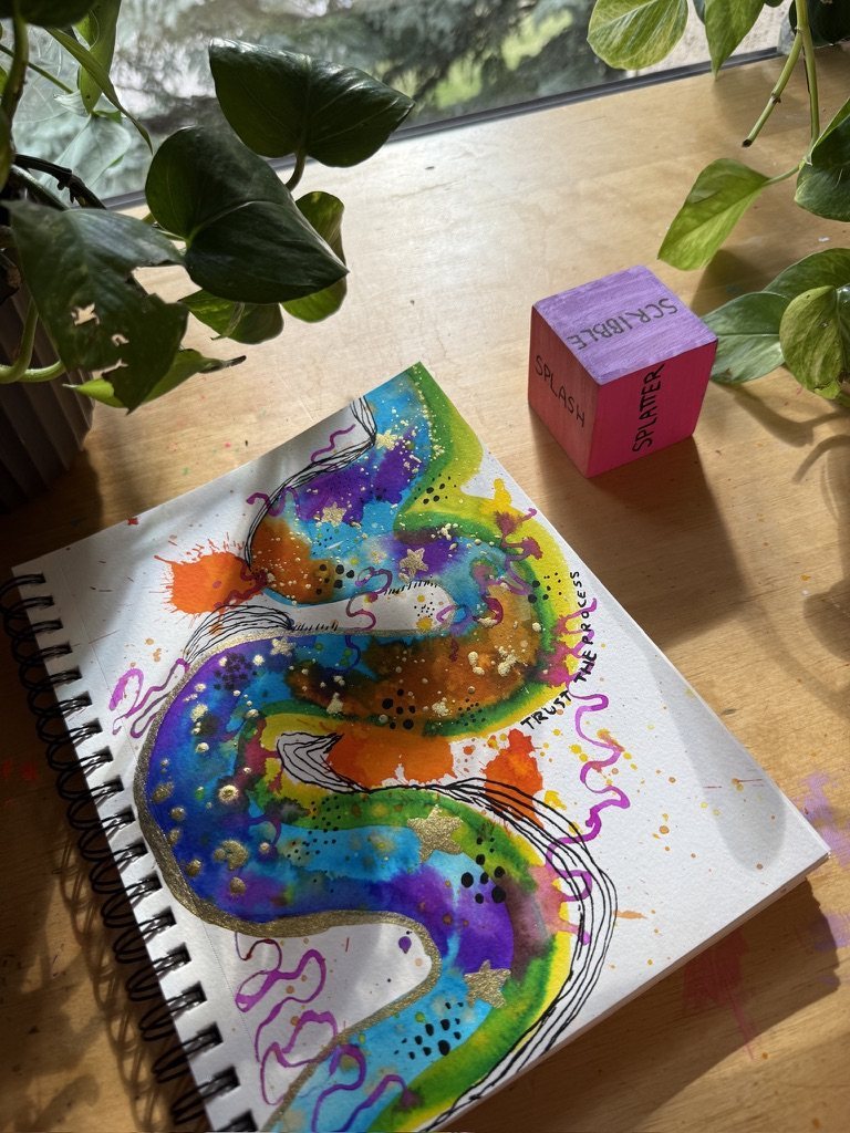 The watercolour sketchbook open on a sunny wooden desk with a pink painted cube and a trailing plant