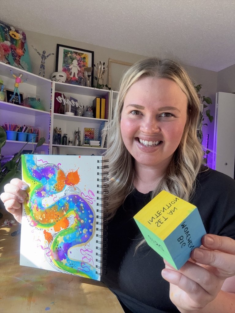 Ashleigh smiling, holding a watercolour sketchbook page and a wooden cube with prompt words painted on its sides
