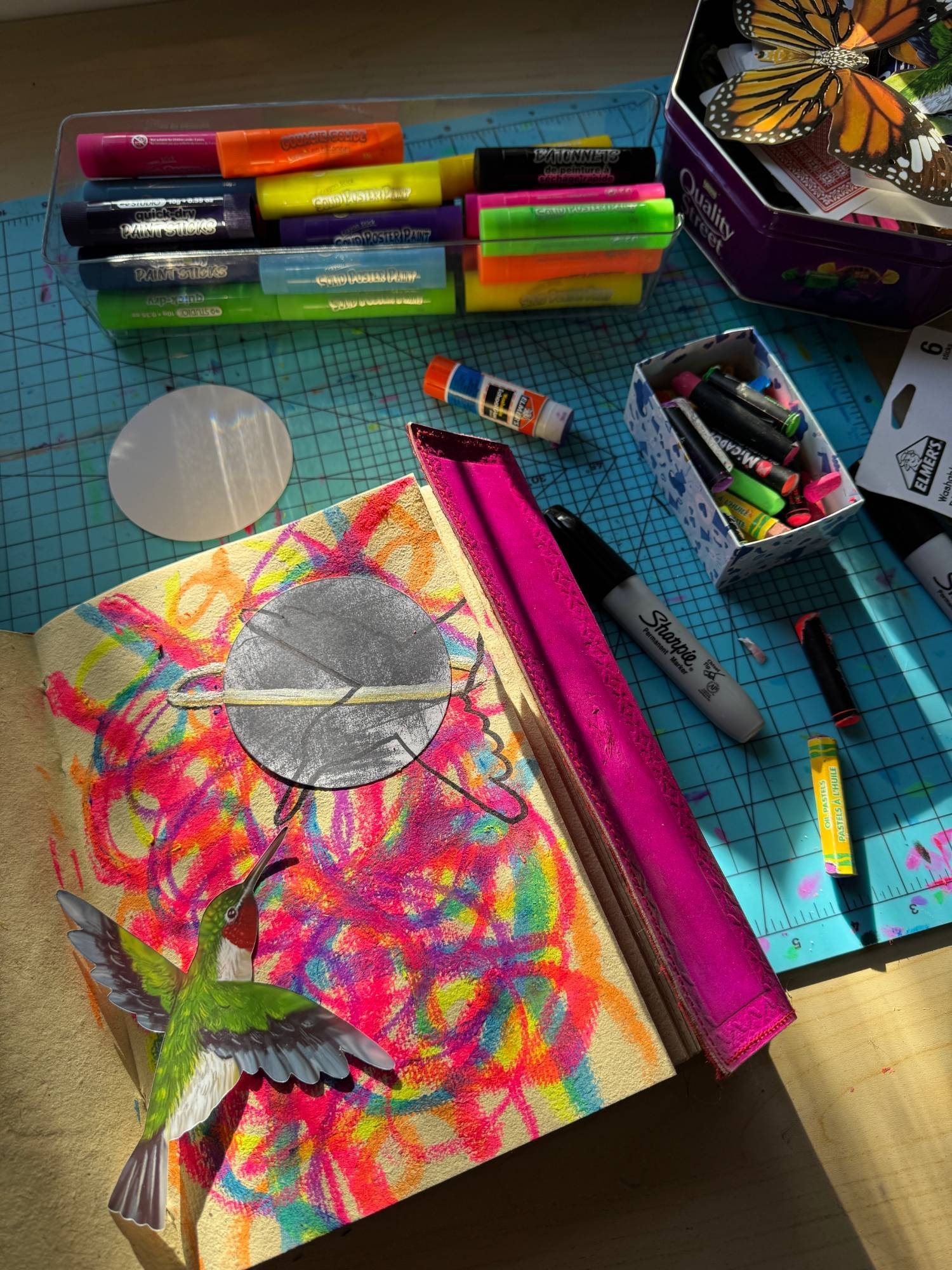 A sketchbook page of rainbow oil pastel scribbles with a paper hummingbird and a silver planet collaged on top