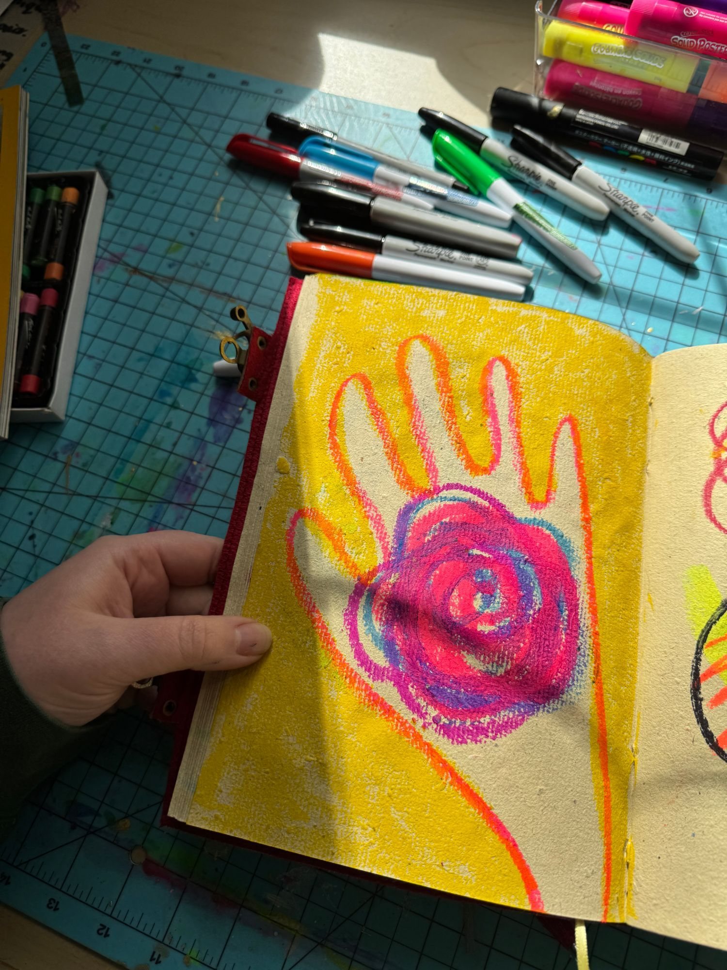 A hand holding open a sketchbook page with a bright yellow background, an orange hand outline and a pink spiral in oil pastel