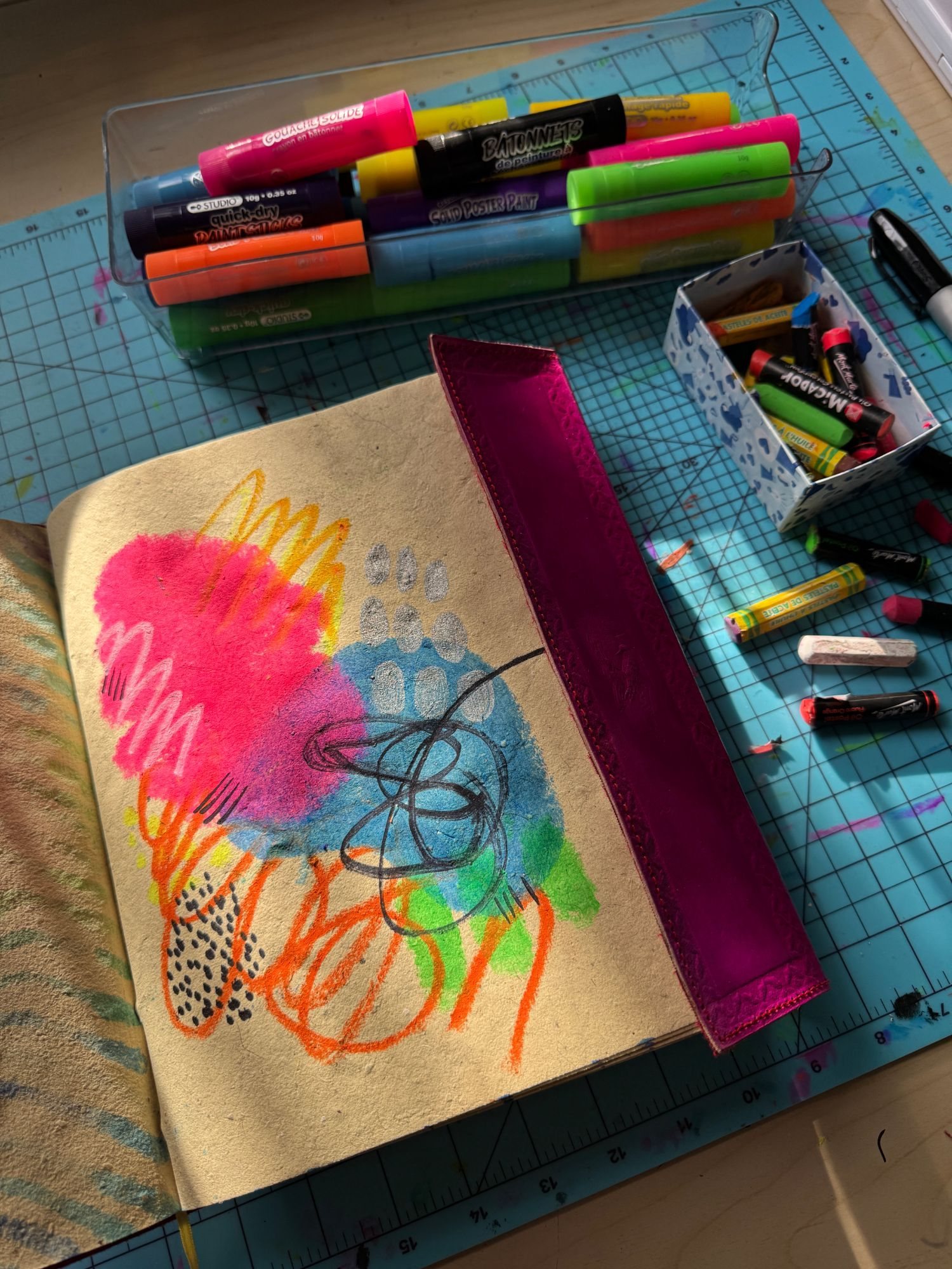 A pink and blue sketchbook page with orange scribbles, beside paint sticks and oil pastels, all laid out and ready to use