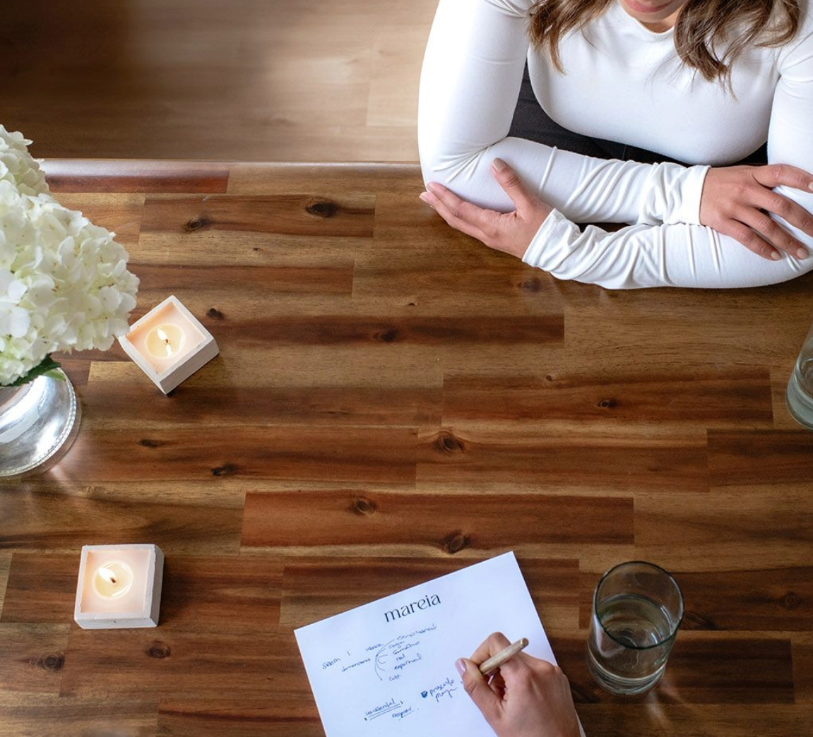 Overhead view of two people at a wooden table during a coaching session, one writing on a mareia notepad