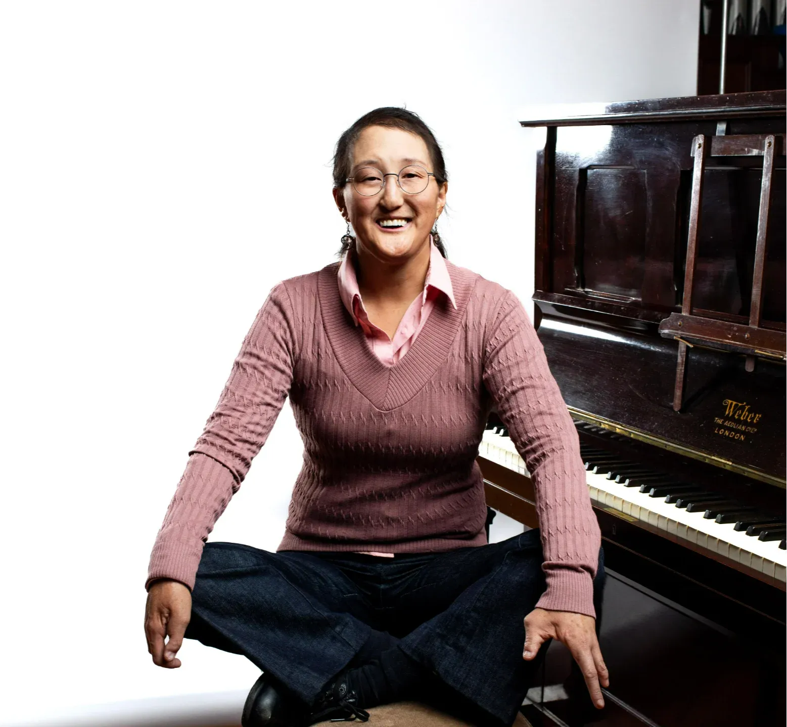 Online piano teacher Camille sits cross-legged on a piano bench next to an upright piano