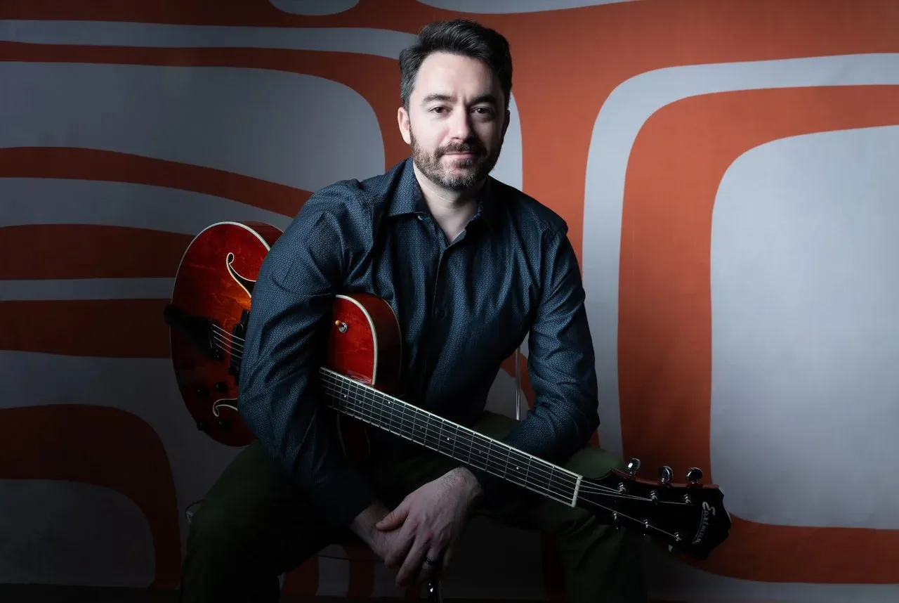 Ben Kunkel, an online guitar teacher, smiles while holding his classical guitar