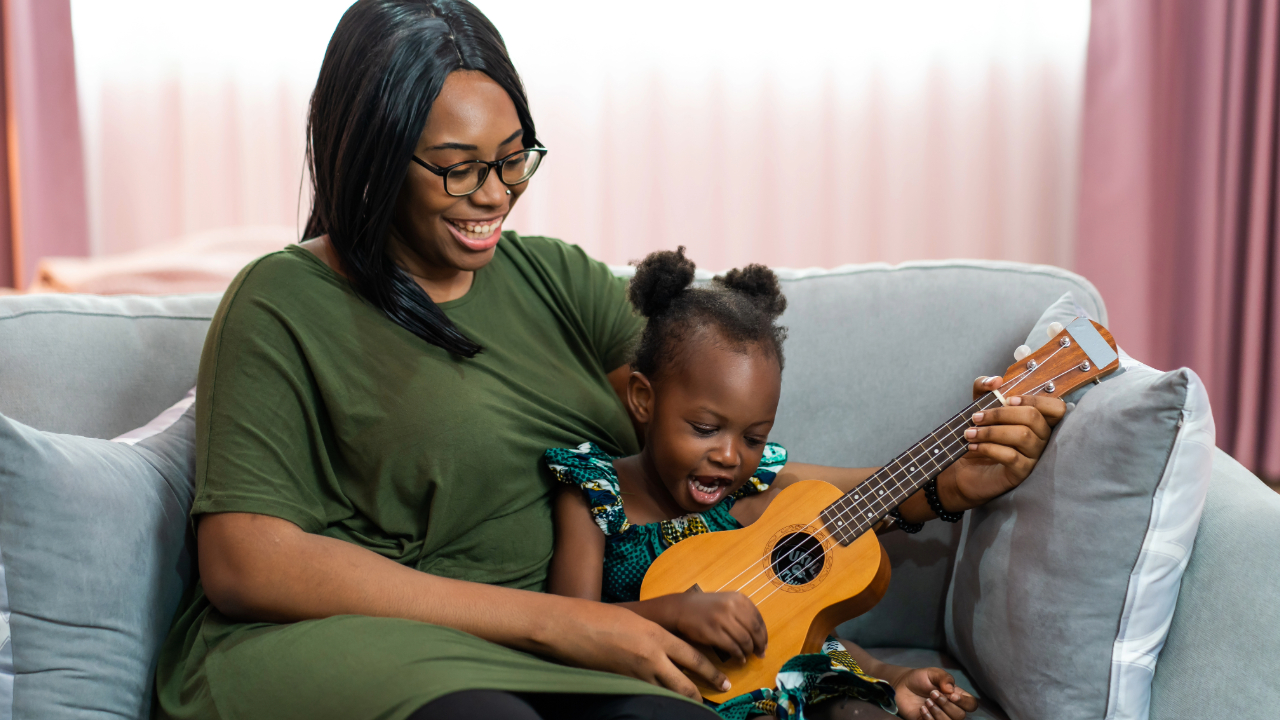 A mother and young daughter smile and sing together while the girl plays a ukulele at home.