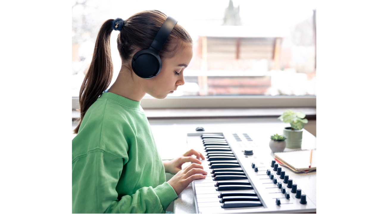 A young girl wearing headphones plays a keyboard at a home workspace, focused and engaged in her music lesson.