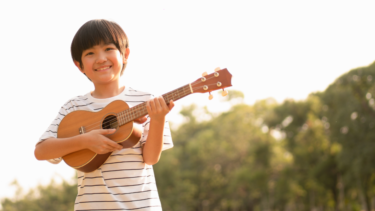 A young boy smiles while playing the ukulele outdoors at sunset.
