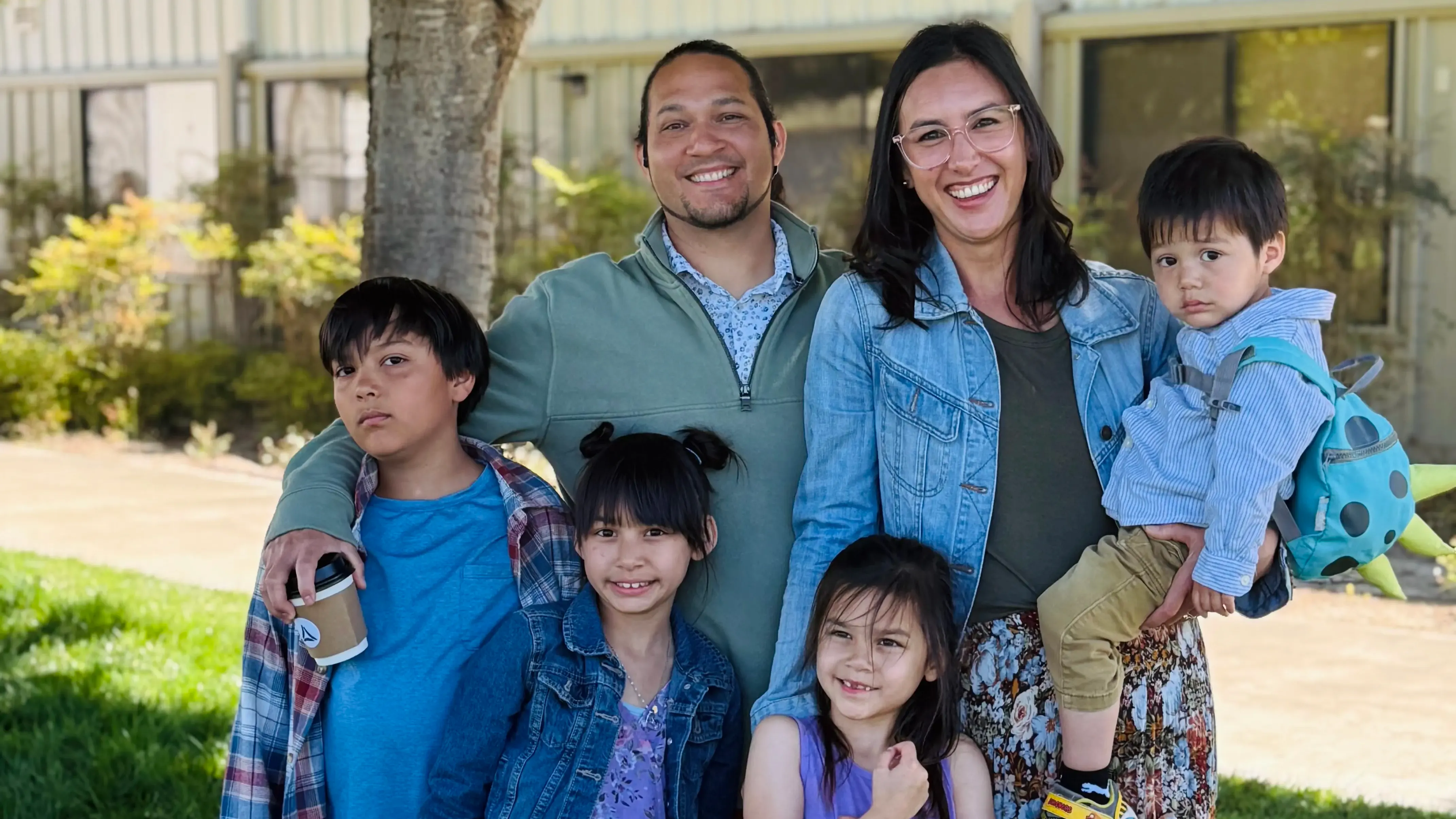 A family of six — two parents and four children — smiles together outdoors under a tree.