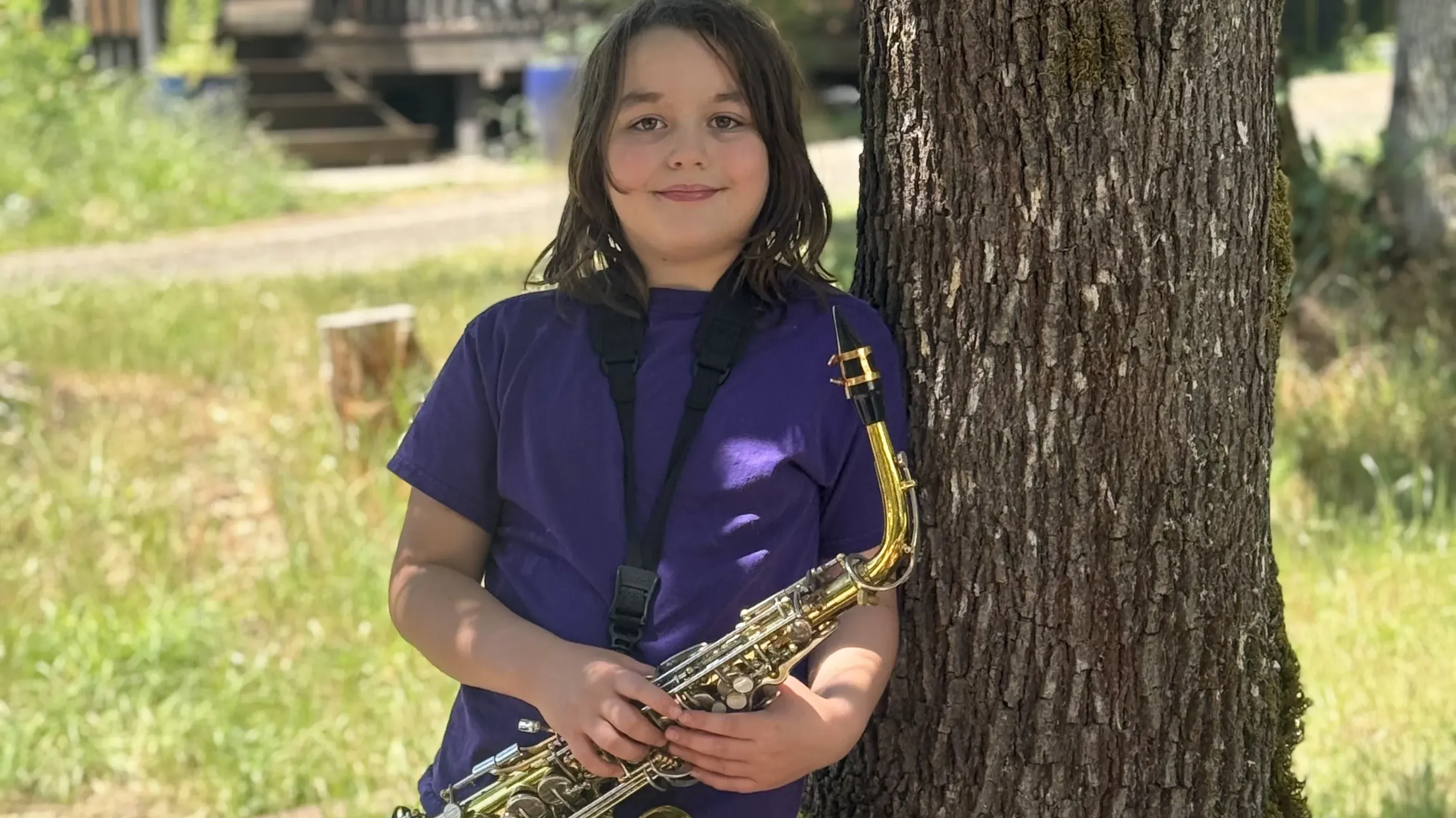 A boy stands under a tree holding a saxophone outdoors.