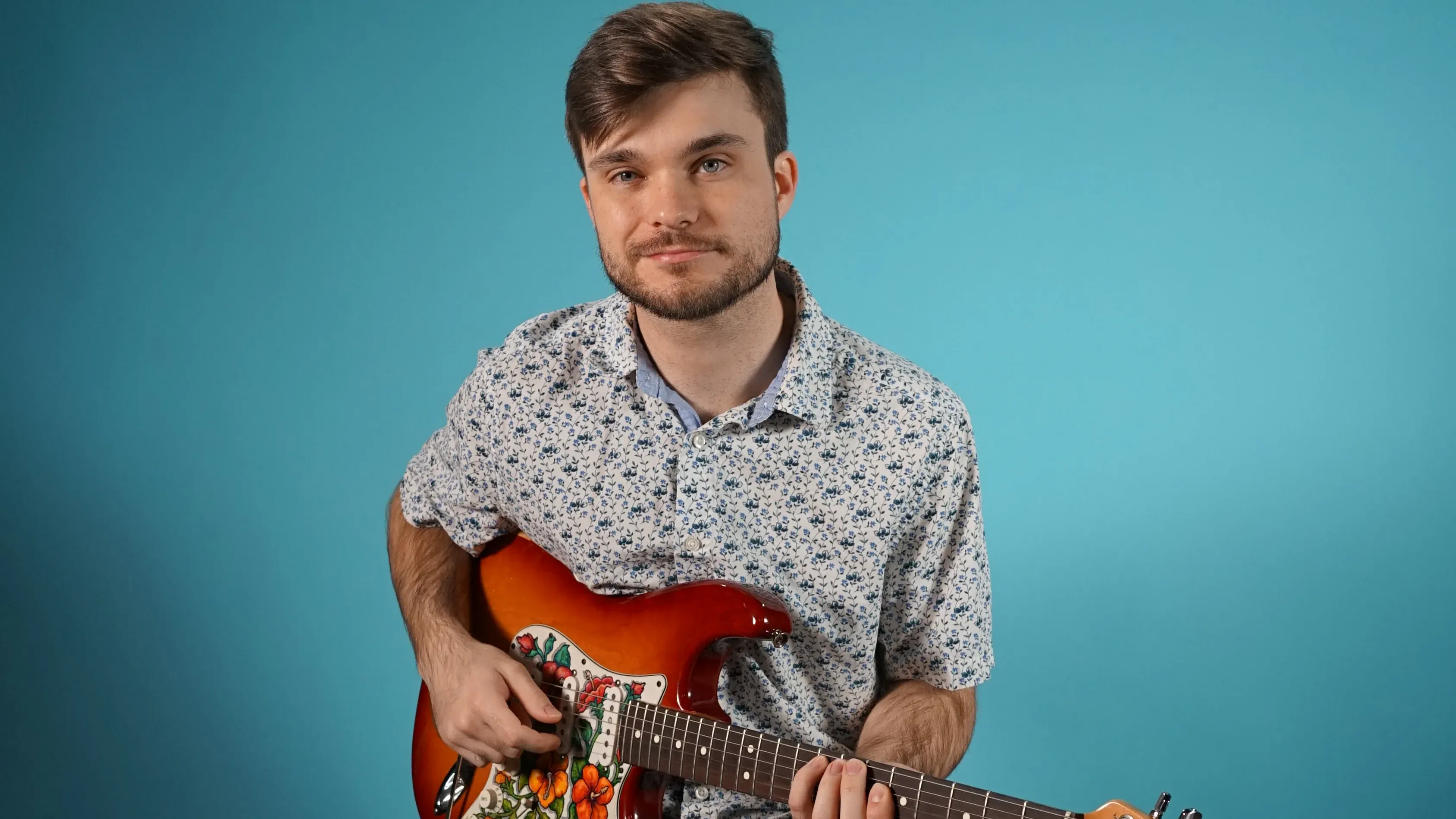 Maestro Music bass teacher, Luke, sits smiling on a stool holding an electric guitar with a blue background
