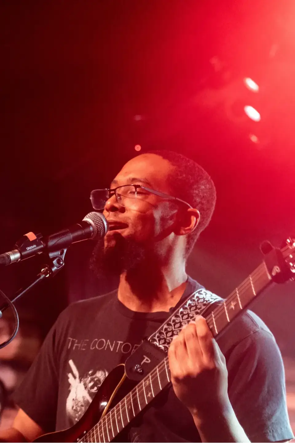 McKinley Hudson, an online piano instructor with Maestro Music, sings into a microphone while holding a guitar on stage