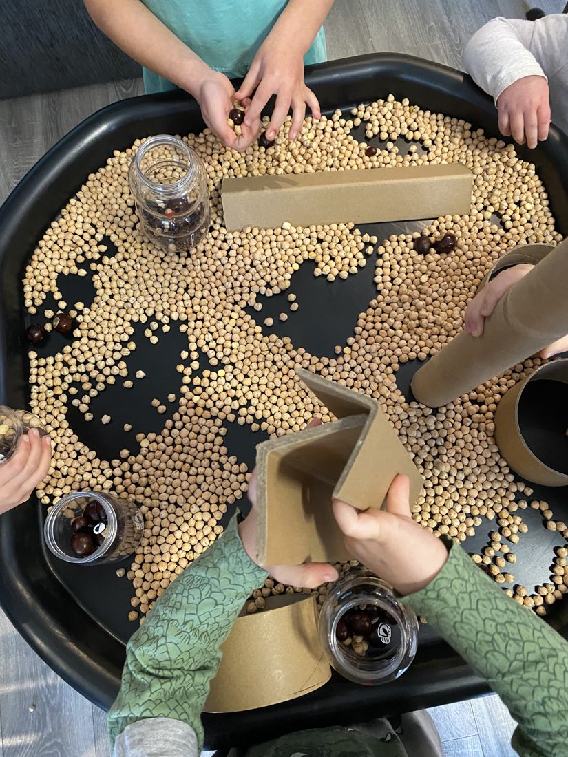 Little hands scooping dried chickpeas and conkers in a black sensory tray with cardboard tubes and jars