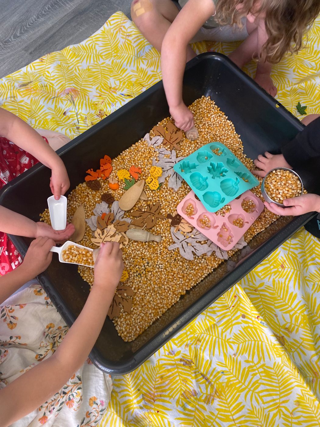 Children scooping popcorn kernels in a fall sensory bin with felt leaves, wooden scoops and silicone moulds