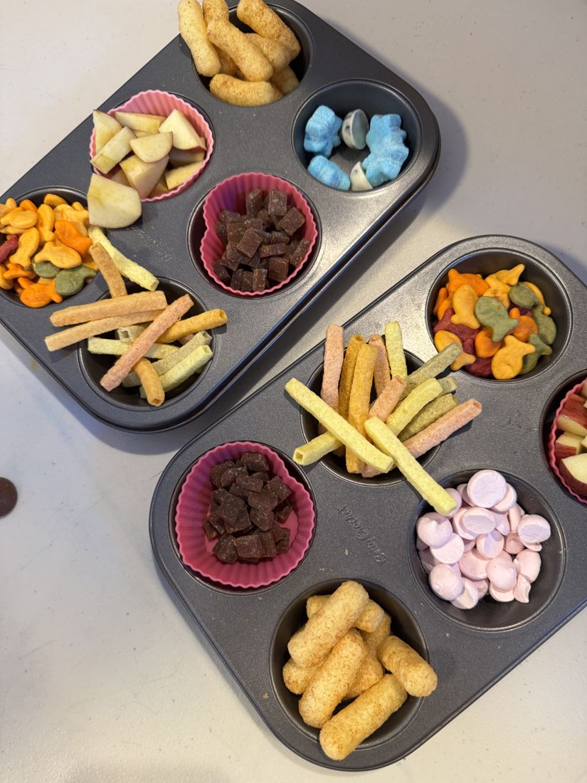 Two muffin tins filled with colourful snacks: apple slices, crackers, veggie straws and fruit bites