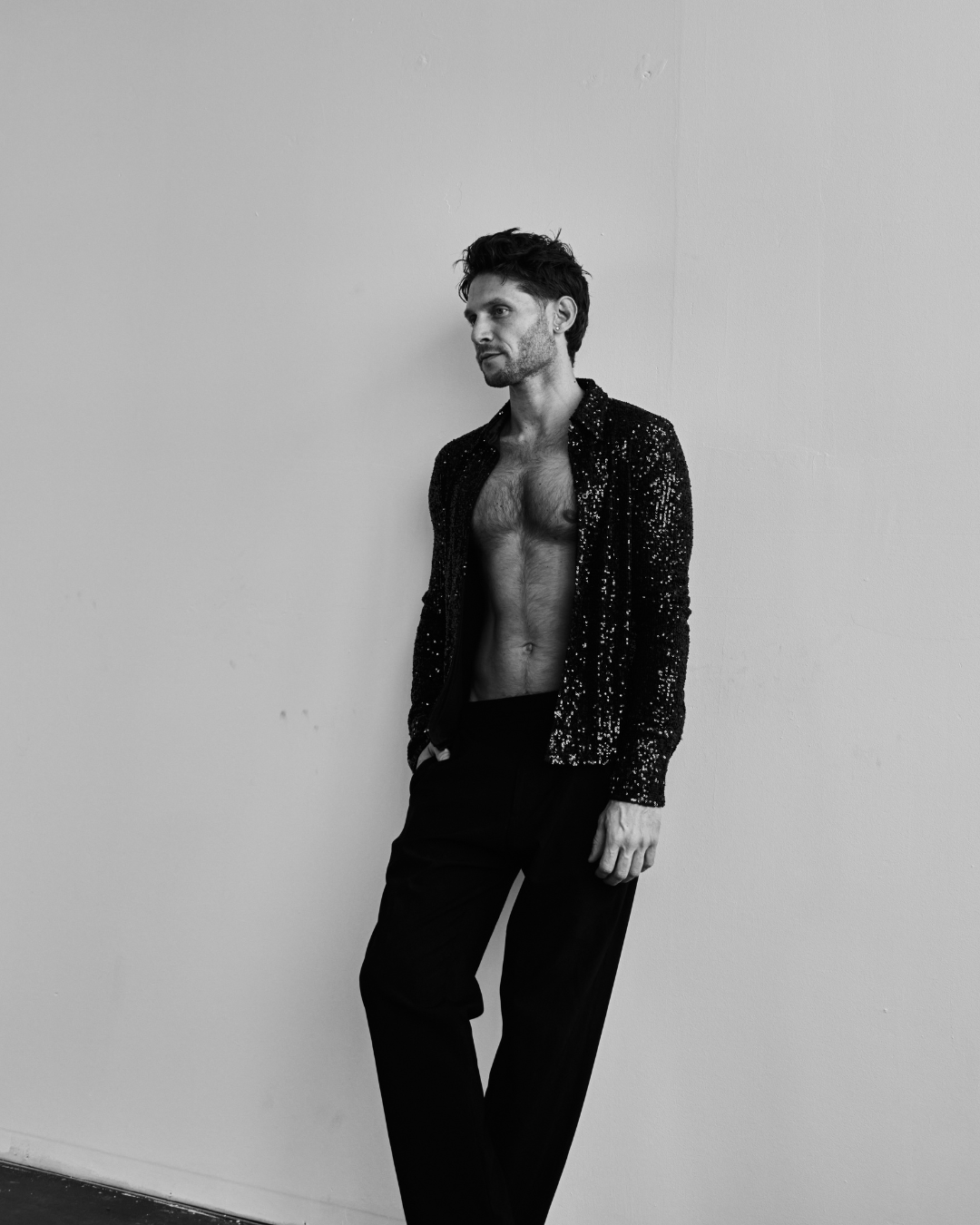 Alessandro leaning on a white wall in a sequin shirt, looking away