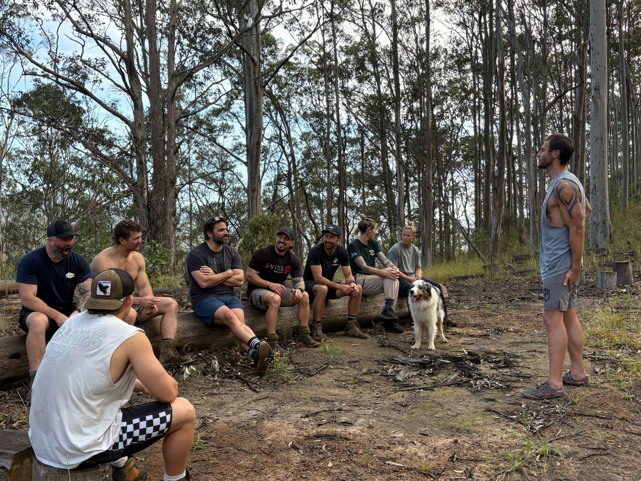 Veterans sitting on a log in the bush, talking with a man standing in front of them, a dog at their feet
