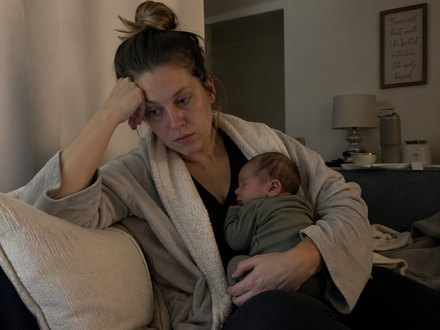 Tired postpartum mother sitting on a couch in soft, dim lighting cradling a sleeping newborn against her chest. She rests her head on her hand with a weary expression, wearing a robe, in a quiet, lived-in living room setting that conveys exhaustion and early postpartum recovery.