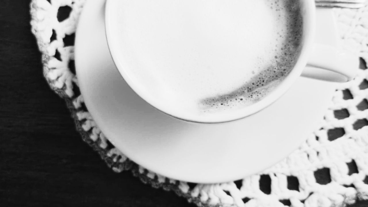 A black and white photo of a frothy coffee in a white cup and saucer on a crocheted lace doily, seen from above.