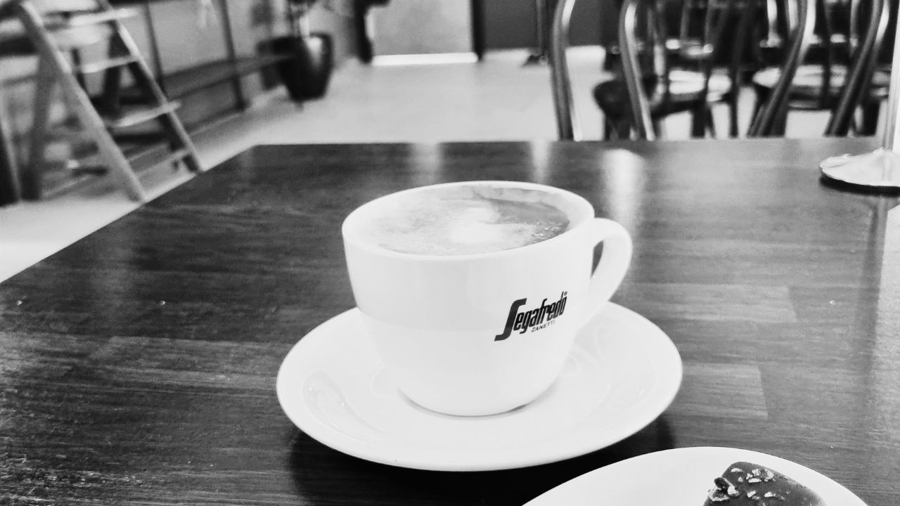 A white coffee cup on a saucer on a dark wooden café table, in black and white.