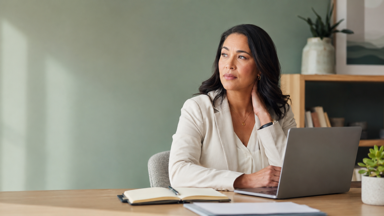 A professional woman sits at her desk appearing calm and capable while gently holding her neck, subtly reflecting hidden stress and burnout.
