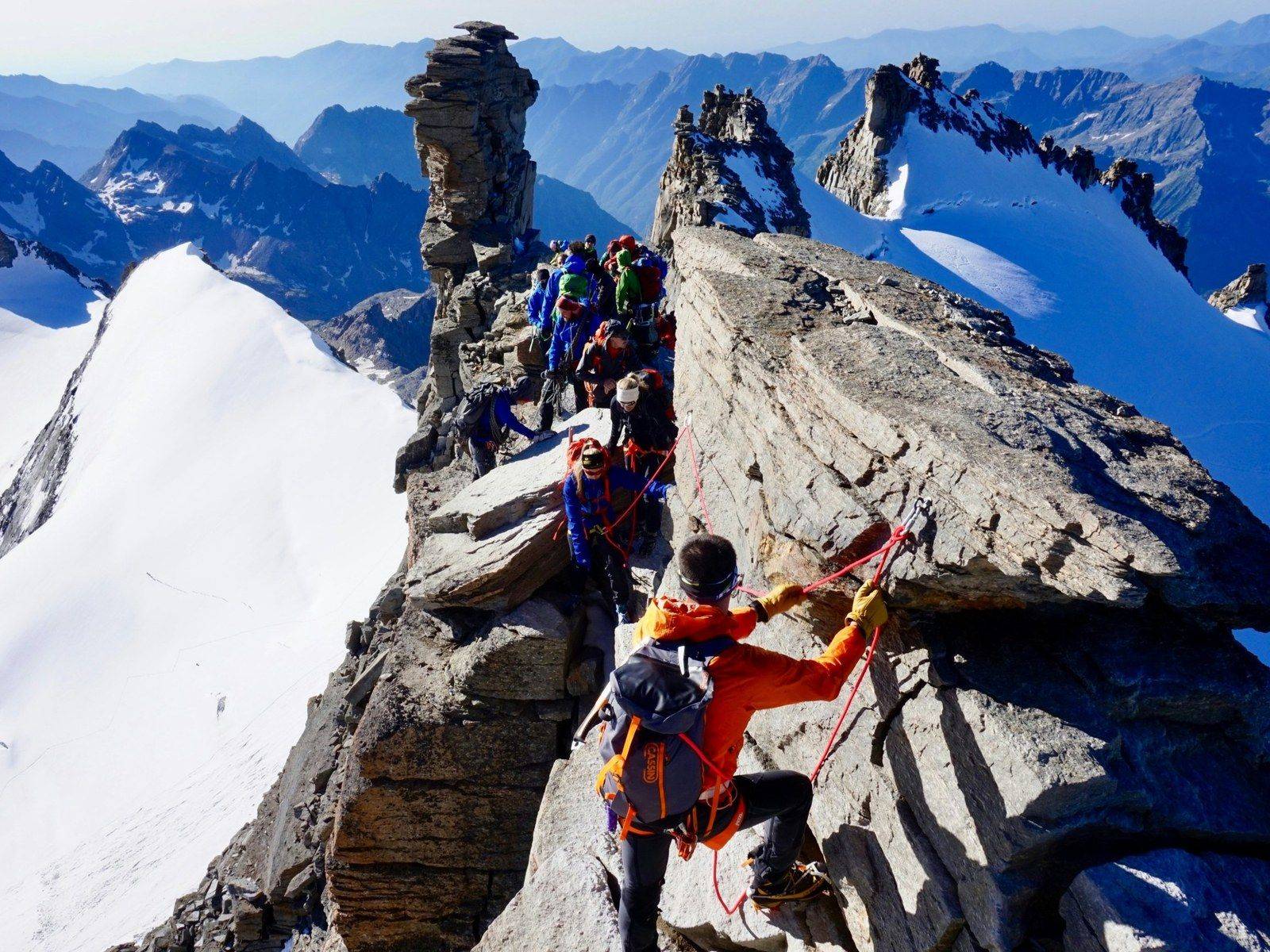 A roped team of climbers moving along a rocky ridge above snow-covered mountains