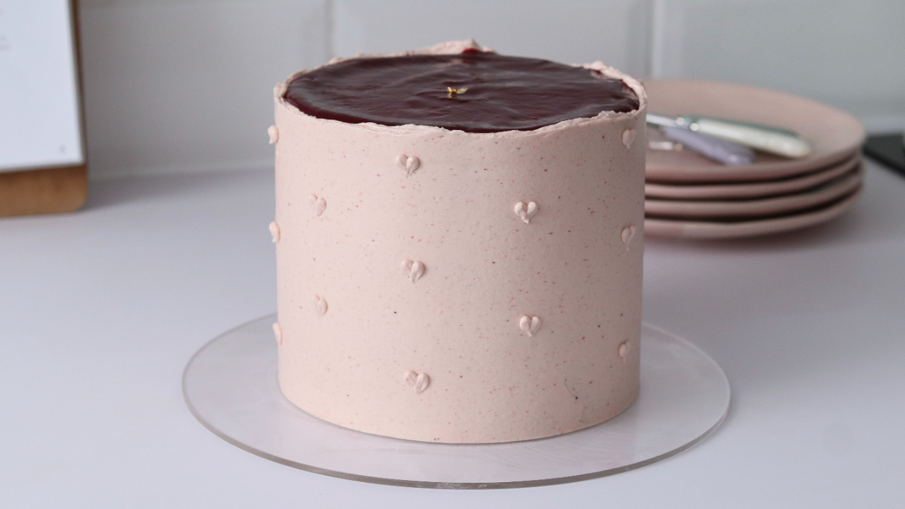 A round layer cake with pale pink Swiss meringue buttercream, decorated with small pearl dots and topped with a smooth dark chocolate ganache drip, displayed on a clear cake board against a white background.