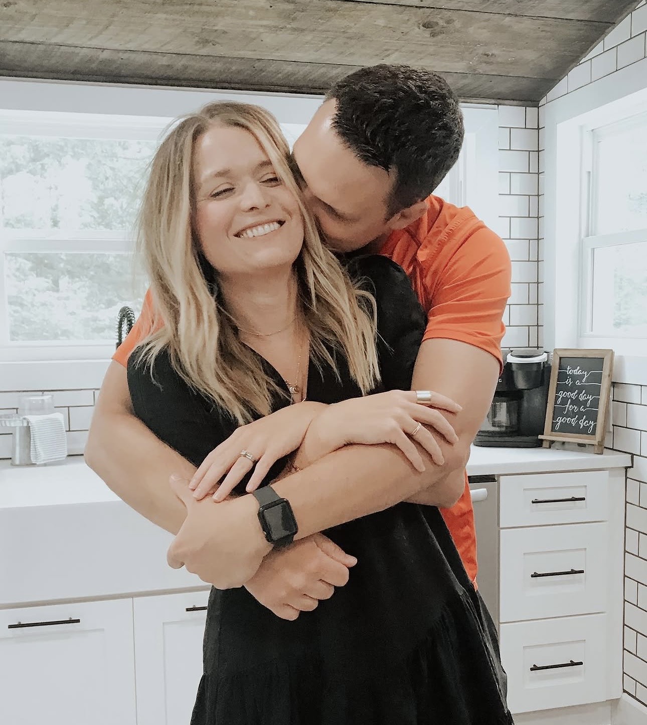 Trae hugging Jess from behind in their kitchen