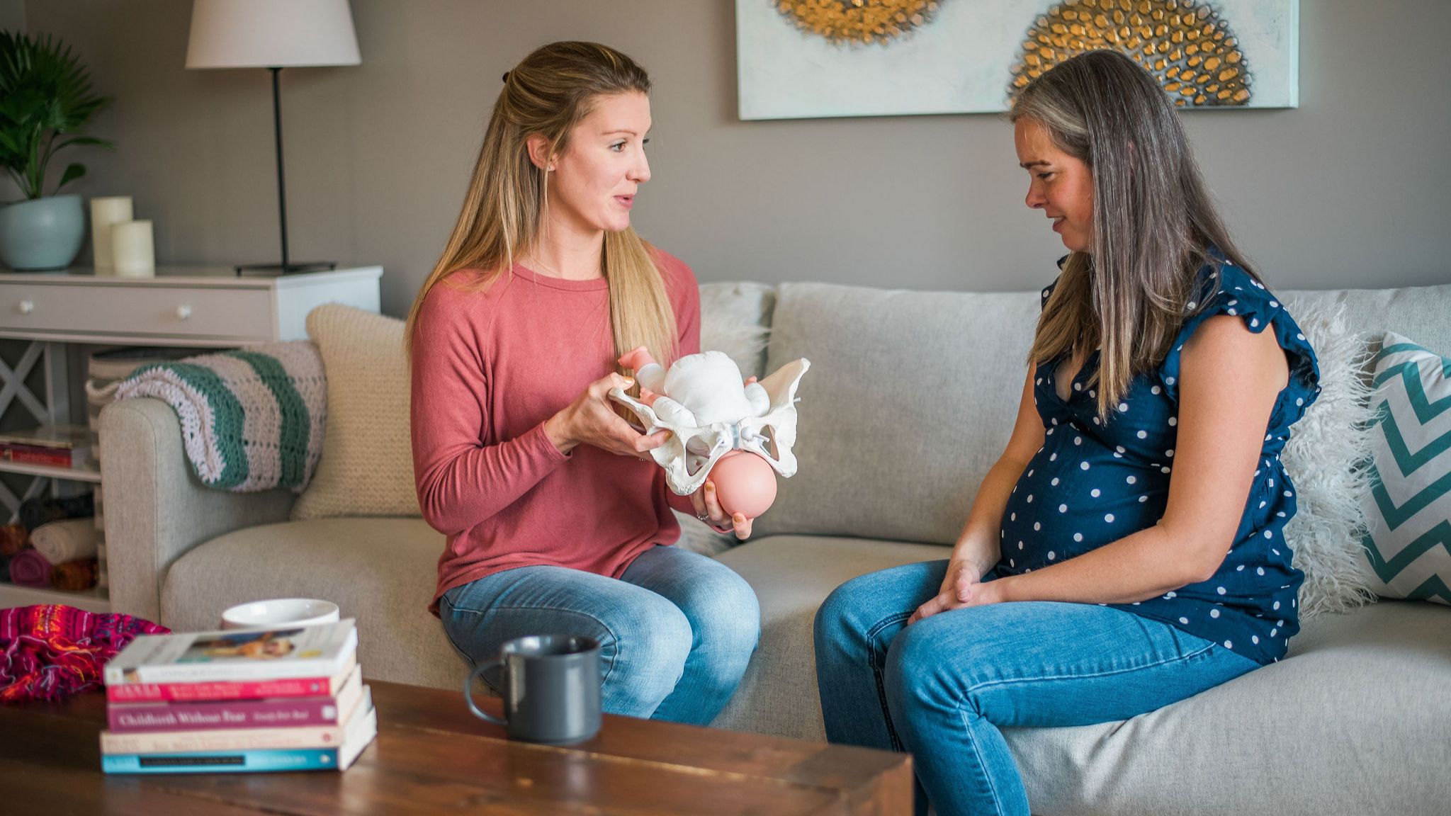 Doula and pregnant woman sitting on a couch facing each other. The doula is using a model pelvis as an educational tool. 