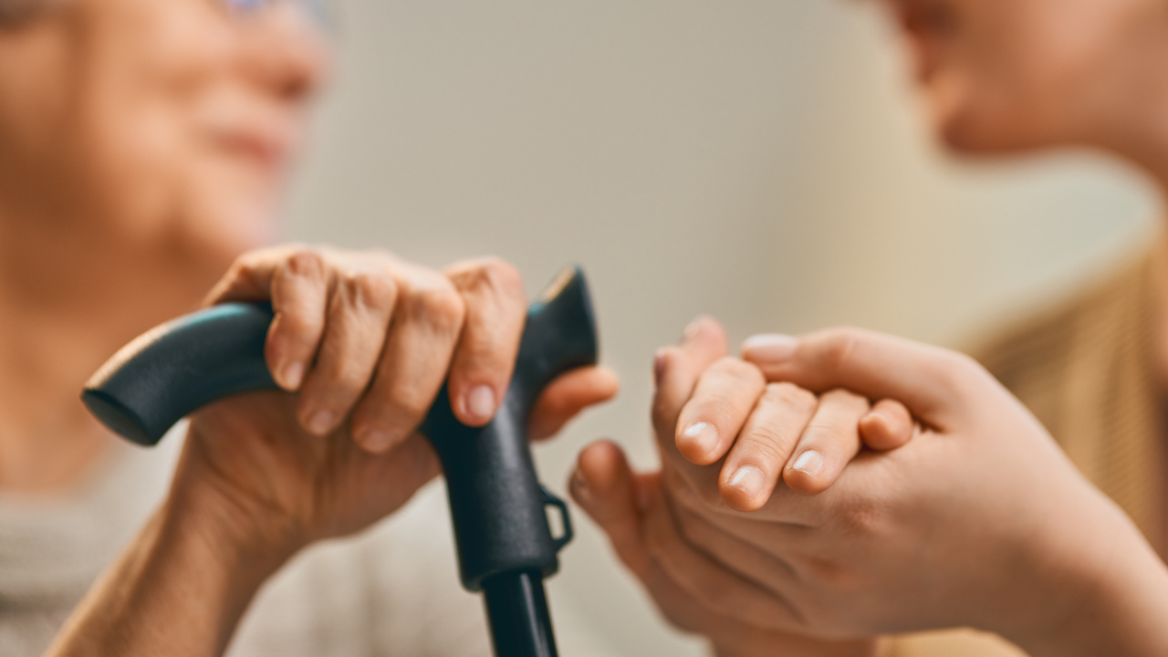 Caregiver gently holding an older adult’s hand while assisting with mobility support and daily elderly care at home.