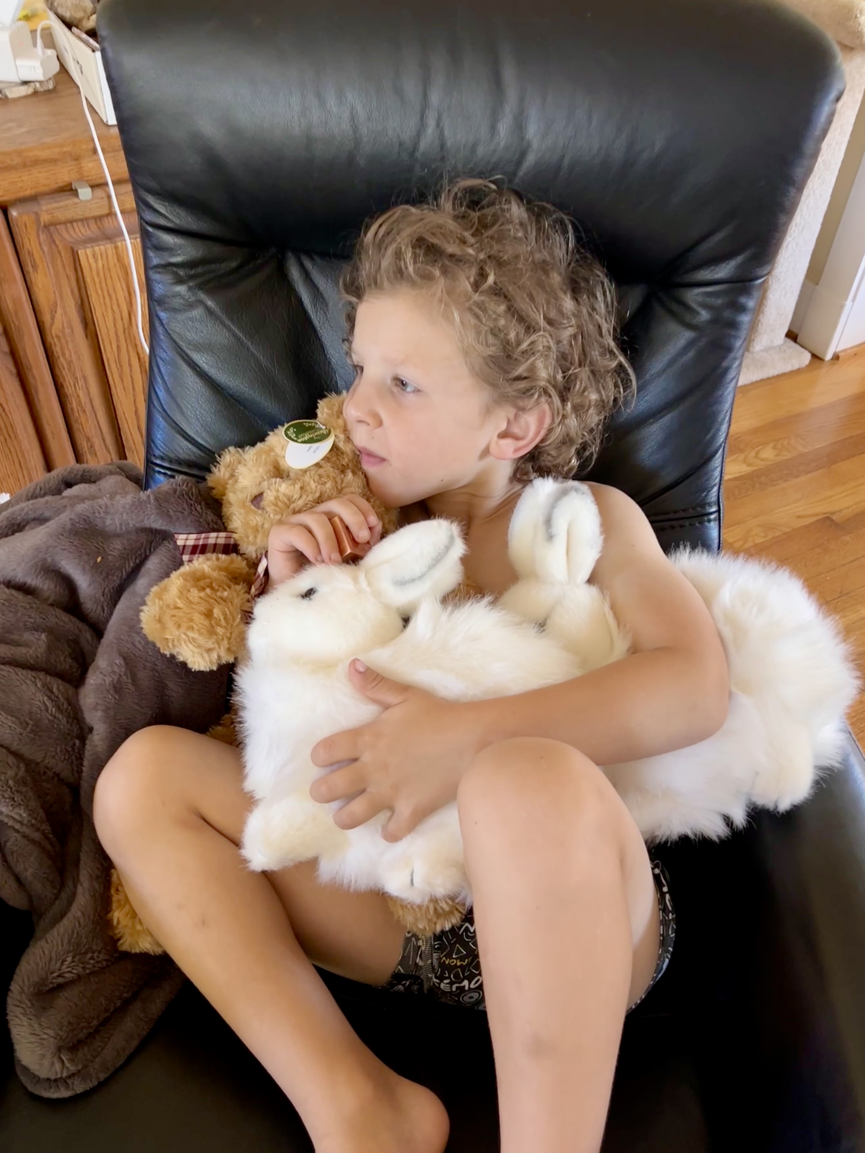 A young boy is sitting in a black recliner watching TV and holding one brown teddy bear and two stuffed white rabbits.
