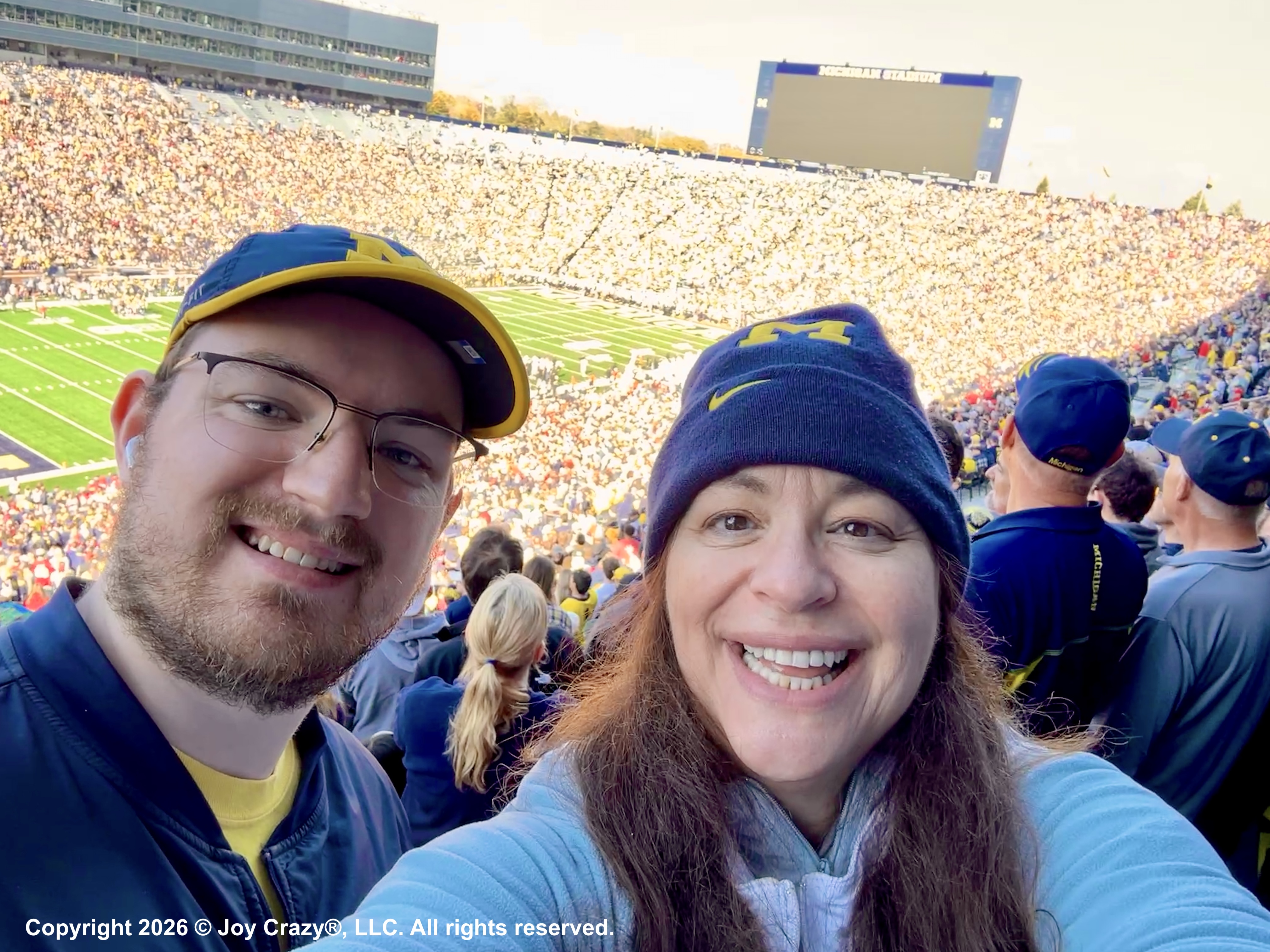 A man and woman smiling at the camera, behind them is a football field and large stadium filled with fans and 
