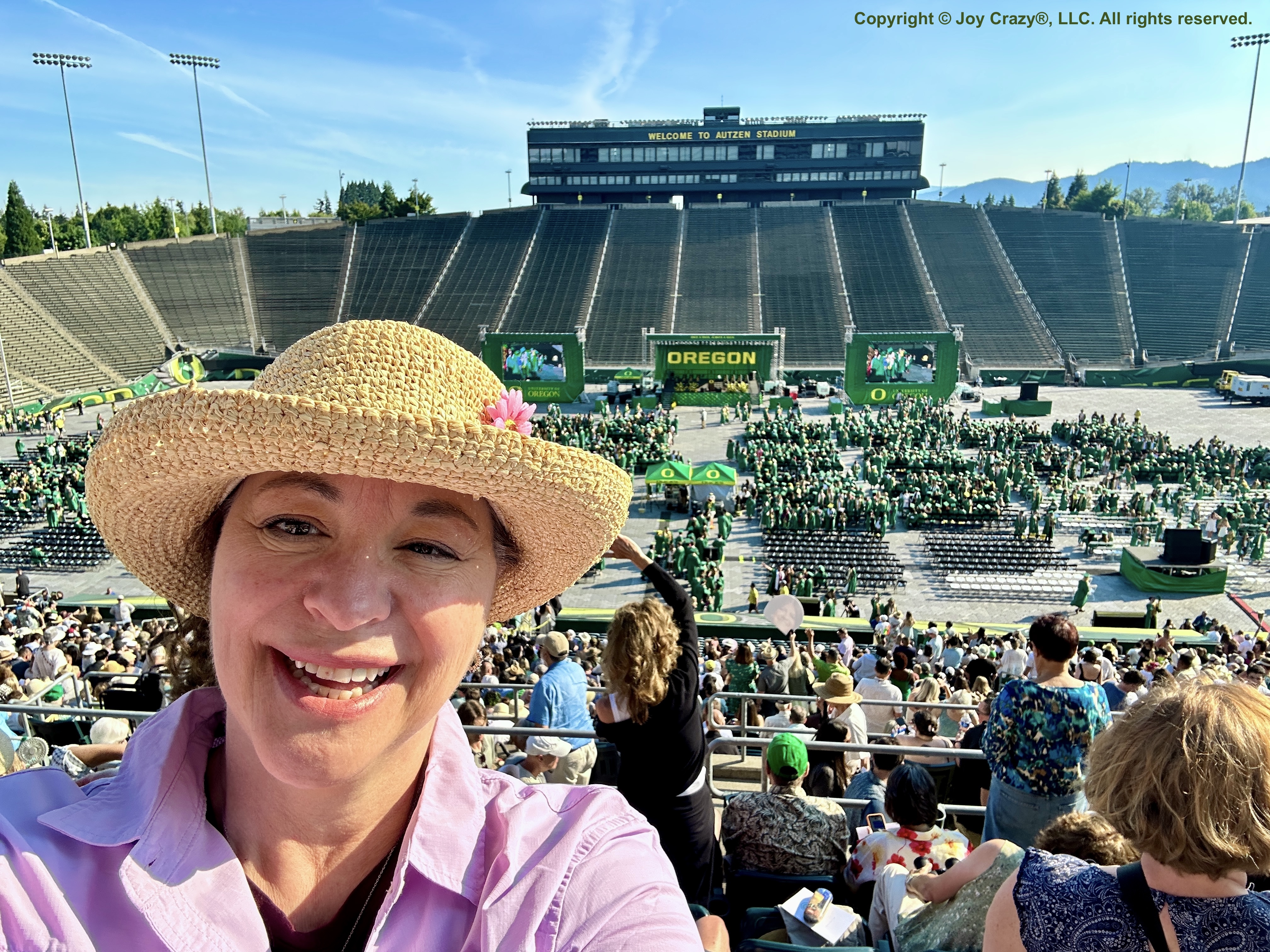 Shannon in the stands of Autzen Stadium where commencment ceremony is about to begin. The graduates wear green robes and sit in chairs facing the speakers stage.