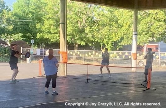 Joy Crazy Pickleball began on an outdoor court. Here four players are under a pavillion playing pickleball on a taped court in the shade.