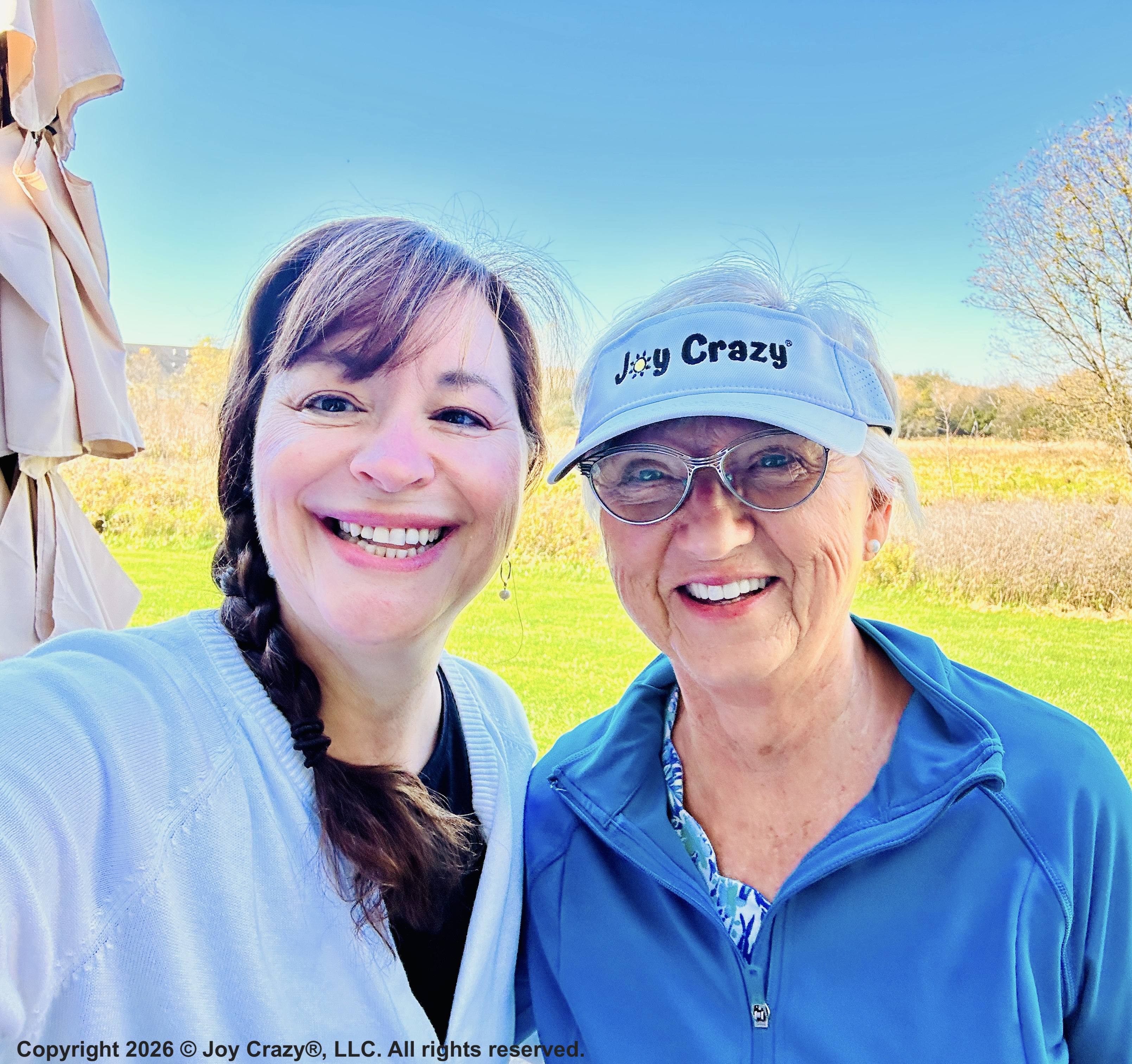 Two ladies smiling at the camera on a sunny day, one wears a visor that says, "Joy Crazy" and behind them is a green marsh.