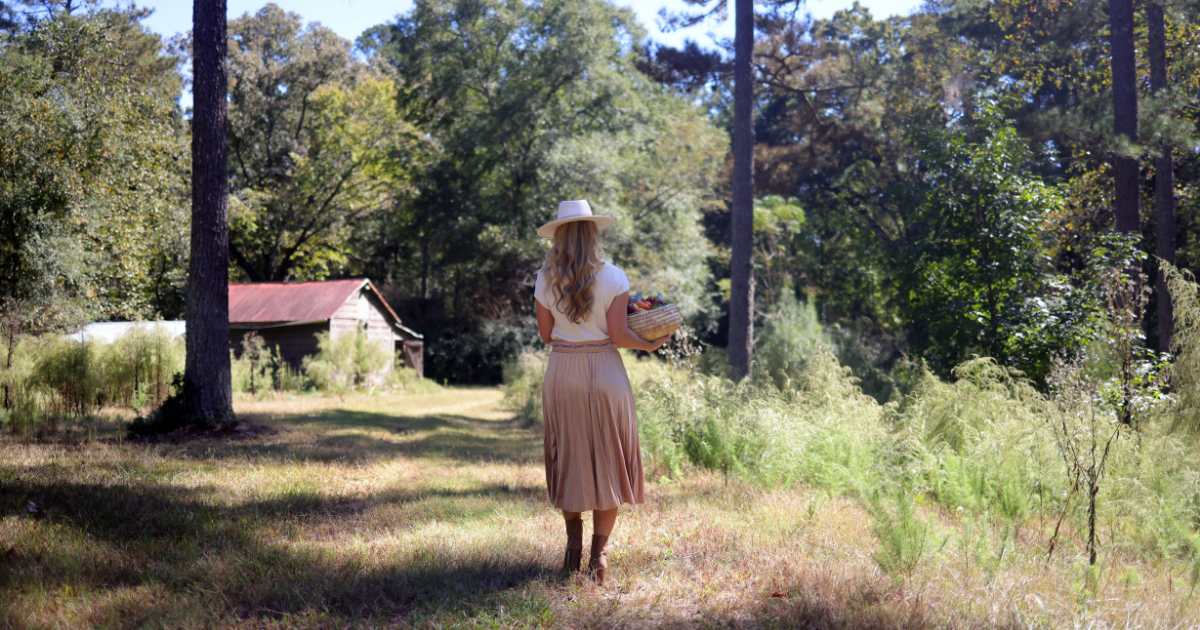 Chef Tiesha walking toward a barn in a wide brim fedora, white short sleeve blouse, and pleated A-line velvet skirt with boots, carrying a basket of vegetables
